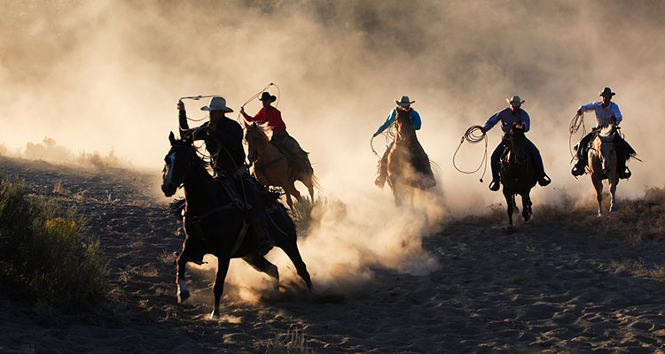 Five cowboys on horseback traveling through a dusty field at dusk.