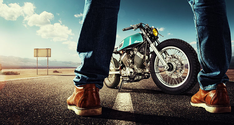 A motorcyclist standing in front of his bike on a desert highway with mountains in the background