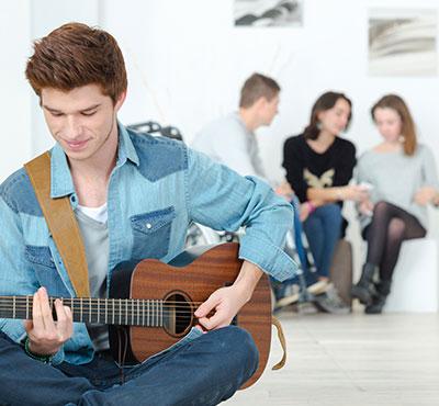 Young man playing a guitar and a group of teens in the backgrounds.