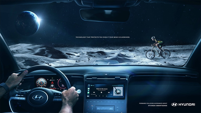 Hyundai brand marketing print advertisement. A hyundai fron inside the car looking at driving on the moon with a cyclist in front.