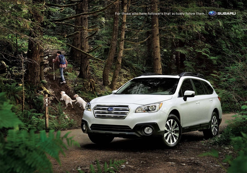 Subaru print advertisement. A subaru outback SUV in a forest with a man going for a hike and 2 golden retriever dogs following him.